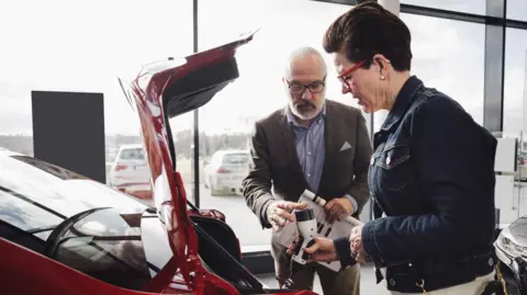 Salesman in a car showroom shows plug connected to an electric vehicle to a female customer 