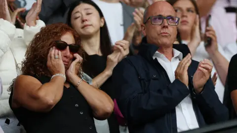 PA Media Jota's mother and father are wearing dark clothing and are both emotional during the tribute. Isabel has her hands on her faces and is crying behind sunglasses. Joaquim has a white short on under a jacket and glasses. He is clapping.
