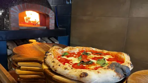 A freshly cooked sourdough pizza, topped with tomato sauce, pepperoni, fresh basil and cheese, is brought out of a lit pizza oven on a wooden board