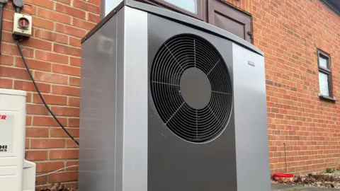 A large grey heat pump outside a brick building in front of a window with a brown windowsill.