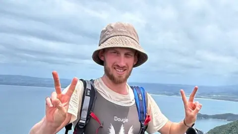 Dougie Haynes is doing a V peace symbol with both hands. He is standing in front of a point where ocean meets land, with land visible on the other side of the water.