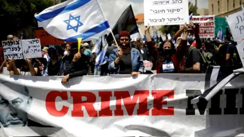 Reuters Protesters wave an Israeli flag and hold placards as they stand behind a banner reading "Crime Minister" before the start of Benjamin Netanyahu's corruption trial in Jerusalem (24 May 2020)