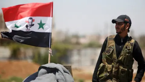 Reuters A Syrian army soldier stands next to a Syrian flag in Umm al-Mayazen, in the Deraa countryside, Syria (10 July 2018)