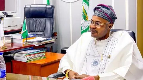 Prince Adeniyi Adeyemi Matthew, PFIPC's director general, sits in an office. He is wearing a white gown, glasses and a grey and pink cap