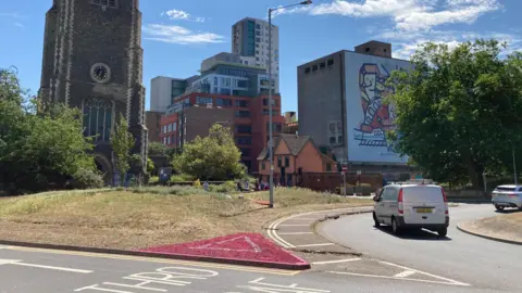 Andrew Woodger/BBC A grass verge off a roundabout is painted pink in the shape of a triangle with a white triangle in the middle of it. Beyond the roundabout are a church, modern high-rise buildings and an old industrial building with a mural on it.