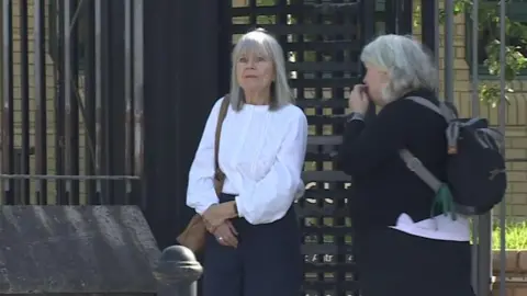 BBC Two women leaving a black turntable style gate. The woman on the left is wearing a white blouse and navy trousers. She has a brown bag on her left shoulder and has straight grey hair to her shoulders and a fringe. The other woman is standing side on and has grey short hair and is all in black. Her face is obscured. She has a black backpack on.