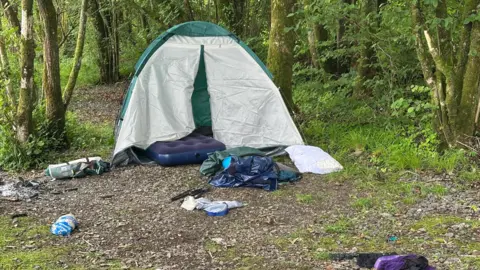 A tent surrounded by litter in picturesque woodland in the Lake District. There is a lot of rubbish and discarded camping equipment. 