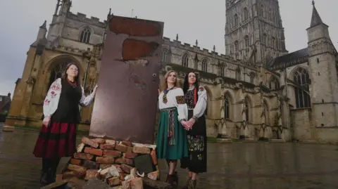 Paul Nicholls Photography Three women, wearing traditional Ukrainian dresses, stand around an art installation – a scorched and ripped red front door. Gloucester Cathedral can be seen behind them on a grey day.