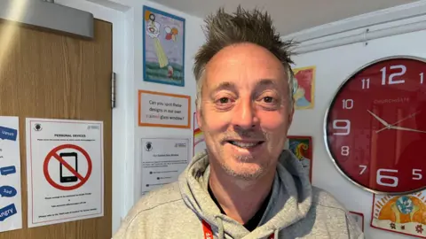 Ashley Scott standing in front of a wall with a red clock to the right and posters to the left. He has spiked up grey hair and stubble, and is wearing a grey hoodie.