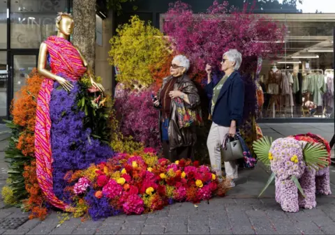 Two ladies look up an a floral design of gold coloured statue of woman in a sari next to a elephant made of pink flowers