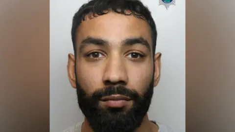 A custody image of Zohaib Mehmood. He has black hair and a black beard and is looking at the camera. 