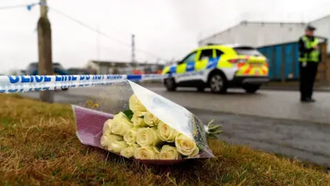 BBC A bouquet of yellow roses placed on the ground with a police cordon in the background