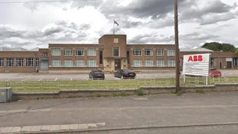 The front of the Stonefield Works in Stone which is a long brown brick building with many windows, which have white window frames. In front is green grass and a car park with three cars on it. 