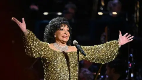 Getty Images Gold sparkly dressed Dame Shirley Bassey grinning after a performance, she has curled black hair and has her hands outstretched 