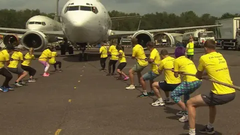 A team of people split in two are pulling an airplane by two ropes. They are dressed in yellow tops and facing away from the camera. It's sunny.