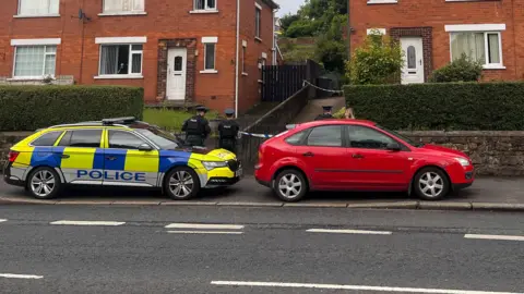 A police car and a red unknown car are parked outside two red bricked houses. A cordon is over one