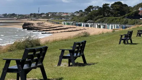 Andrew P The beach and some beach huts in the distance, with waves breaking on the shore. In the foreground there are a row of benches a few feet apart, sat on a grassy field looking out to the ocean.