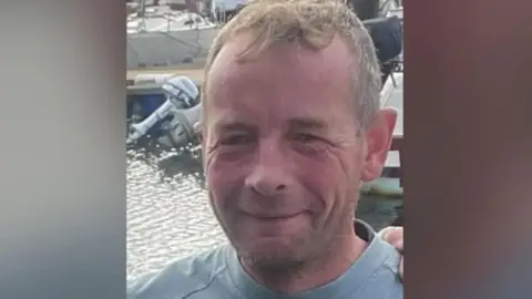 Lee Scott, a man with short strawberry blonde hair smiles in front of the harbour.