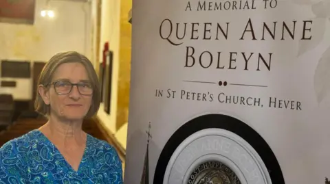 A woman stands in front of a sign that says 'a memorial to Queen Anne Boleyn'.