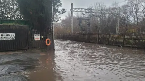 A flooded road, next to a railway line. There is an orange flotation ring on a fence alongside the flooded road. There are trees and fences in the distance.
