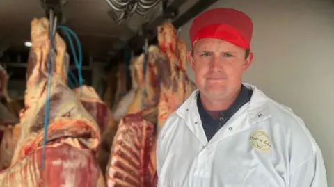 A man in a large white coat and a red hat stood in front of butchered meat hanging from a ceiling