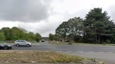 Google Road junction off a major dual carriageway road. There is a crossing point to the right of the photo where eastbound traffic can cross in front of westbound traffic to enter into the Standens Barns area. Two cars are visible to the left on the main road and one has just turned into the side road. There are trees on either side of the road.