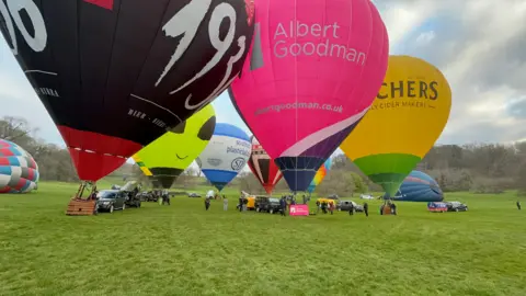 12 hot air balloons being inflated in a field on the Ashton Court Estate in Bristol to launch the Bristol International Balloon Fiesta. People are gathering under the balloons.
