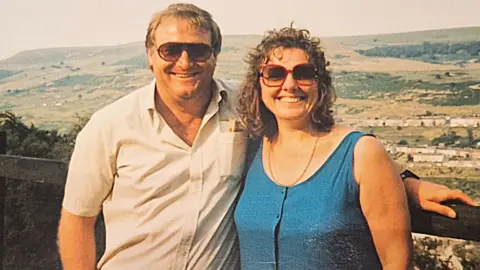 Family photo A photo of Jayne as a younger woman with her late husband. They are posing together at a viewpoint. Jayne has long curly hair and a blue dress and her husband is wearing a beige short-sleeved shirt.