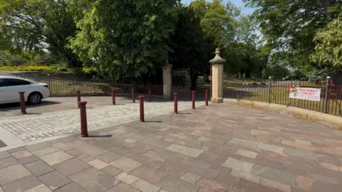 The stone and metal entrance gates of Barnes Park. A banner promoting an upcoming family fun day is hanging on the red fence.
