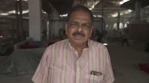 Pritam Roy/BBC Sanjay Jain - wearing a pink and green striped shirt with a phone jutting out of his pocket - stands in his glass bangle factory. Workers can be seen going about their day in the blurred background. 