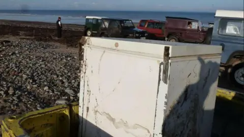 An abandoned freezer on a pebbled beach; in the background lies the sea, a number of parked vehicles and a man standing on the shoreline.