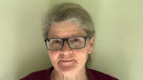 A landscape head shot of GeneWatch UK Director, Dr Helen Wallace. Her head and shoulders are visible and she is standing in front of a magnolia coloured wall. She is wearing a burgundy knitted jumper and has black-framed, rectangular glasses. Her hair is wavy, tied in a ponytail and it grey in parts. 