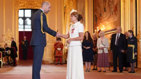 PA Media Ellen Roome, dressed in a white skirt, white blouse, and white hat, smiles as she shakes hands with Prince William during an investiture ceremony. Her MBE can be seen on her chest. There are people standing around, watching, inside an ornate hall at Windsor Castle.