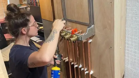 Beth is wearing a blue T-shirt and safety goggles. She is working on a test rig for a heating system that has five copper pipes leading into five different brass outlets. She is holding a blue-and-yellow gas blowtorch for brazing joints in her left hand and soldering wire in her right.