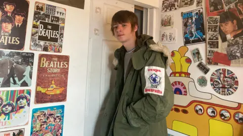 BBC/Louise Fewster Teenage boy with brown hair going over his ears is wearing a green parka jacket with a cream edge on the hood. Travis is stood in front of a white bedroom door with The Beatles posters on the wall to the left and right of him. Behind Travis is a colour photo of a yellow submarine featured on one of The Beatles albums. 