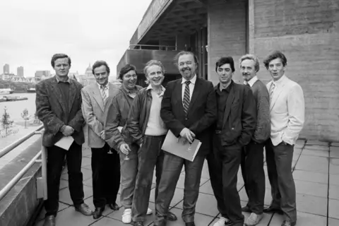 PA Media A line of actors and directors outside the National Theatre's concrete building on the Thames in London. In a row are Sir David Hare, Sir Richard Eyre, Michael Bogdanov, Peter Gill, Sir Peter Hall, Bill Bryden, Edward Petherbridge and Sir Ian McKellen. 