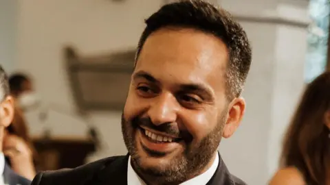 A man with short brown hair and beard who is smiling, looking to one side of the camera. He is wearing a black jacket and white shirt.