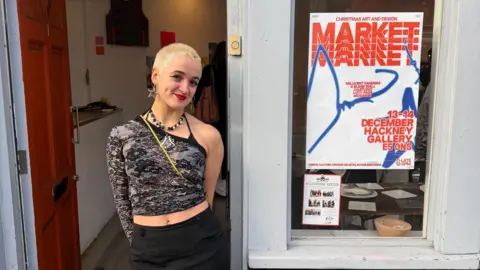 Jess O'Riley A woman with really short blonde hair, wearing an off-the-shoulder top standing outside a fashion studio