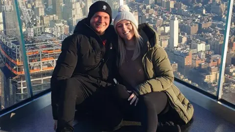 Tasha Quick A man wearing a black hat, coat and trousers and a woman wearing a light grey hat and a green coat with black trousers. They are sitting in front of glass panels with a cityscape visible behind them.