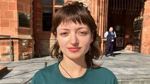 A woman is wearing a green shirt. She has brown hair and multiple face and ear piercings. Behind here is a red building with grey steps and a silver handrail at its front. There is a guard standing outside the entrance. 