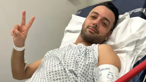 @pouriazeraati Pouria Zeraati lies in what appears to be a hospital gown, and holds up his fingers in a "V" sign.