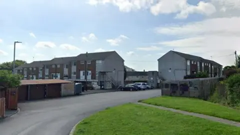 A cul-de-sac in a residential area. Pictured are some closed garages alongside the road, with terraced houses in the background. A grass verge and a footpath is on the other side of the road.