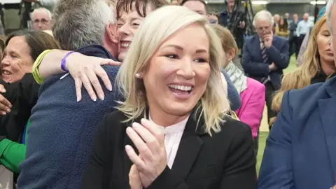 Sinn Féin deputy leader Michelle O'Neill clapping and smiling broadly. She is wearing a black jacket over a white shirt and has medium-length blonde hair