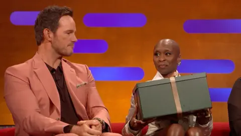 Cynthia Erivo holds a green gift box with a gold ribbon. Chris Pratt, to her right, looks at the box