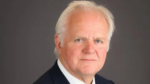 BCP Council Official studio photo of councillor Mike Cox looking at the camera with a serious expression. He has white hair and is wearing a black suit with a white shirt