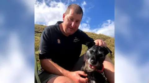 Stephen Hale, wearing a dark Guinness polo shirt, sitting on a hill. He is stroking a wiry, black dog, which is wearing a red collar.