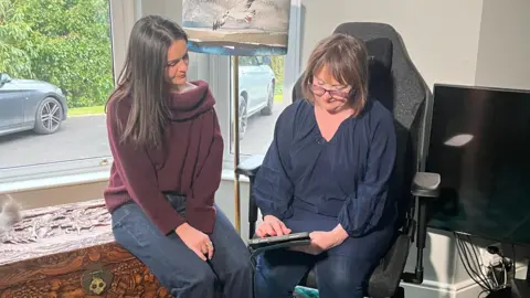 Suzy with her mum Margie. Suzy is sitting on a trunk beside a window and Margie is sitting on a computer chair wearing glasses. Margie is showing Suzy something on an iPad.