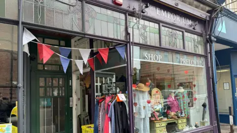 The Emporium shop front is painted deep purple and green. Clothes and furniture can be seen in the display. Red, white and blue bunting hangs over the doorway.