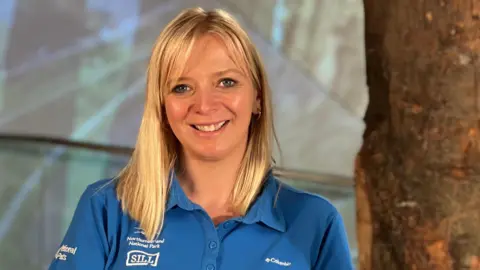 Rosie Thomas smiles at the camera. She has shoulder-length blond hair and is wearing a blue polo shirt, and is standing next to the trunk.