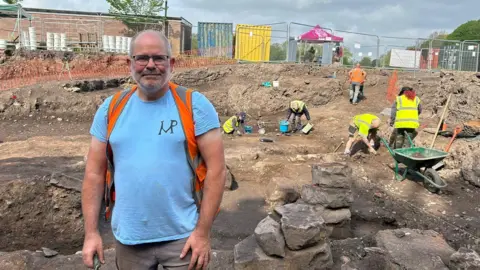 Anna Giecco Frank Giecco has short grey hair at the sides. He is wearing glasses, a blue t-shirt, orange/grey rucksack, beige trousers. He is stood on the dig site with soil and rocks. There are volunteers with PPE on with tools and a green wheelbarrow. There is a temporary metal fence in the background a small brick building and large storage shipping containers.